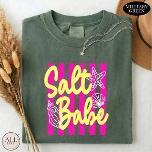 a green t-shirt with a pink and yellow design that says "Salt Babe" on it. The t-shirt is folded and placed on a white surface, with a brown hat and some dried flowers nearby.