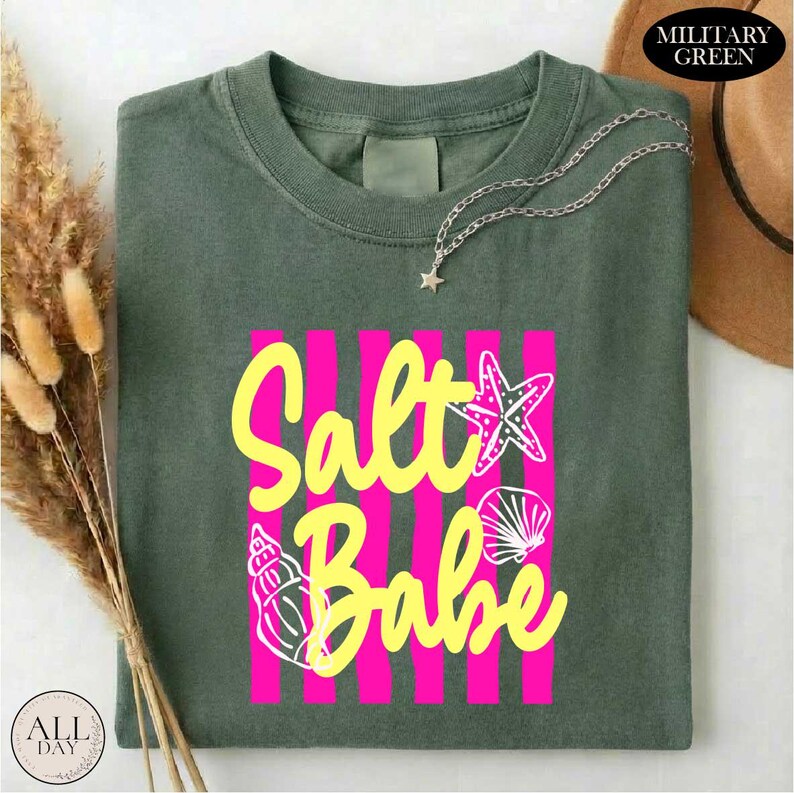a green t-shirt with a pink and yellow design that says "Salt Babe" on it. The t-shirt is folded and placed on a white surface, with a brown hat and some dried flowers nearby.