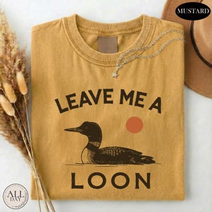 a yellow t-shirt with a graphic design of a duck and the text "LEAVE ME A LON" printed on it.