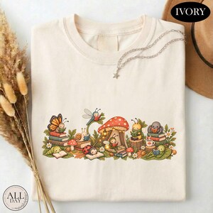 K&ouml;nnte beinhalten: Ein elfenbeinfarbenes T-Shirt mit einer grafischen Illustration verschiedener Insekten, darunter ein Schmetterling, eine Schnecke, ein Marienk&auml;fer und eine Biene, die um einen gro&szlig;en roten Pilz herum B&uuml;cher lesen. Das Design enth&auml;lt gr&uuml;ne Bl&auml;tter.