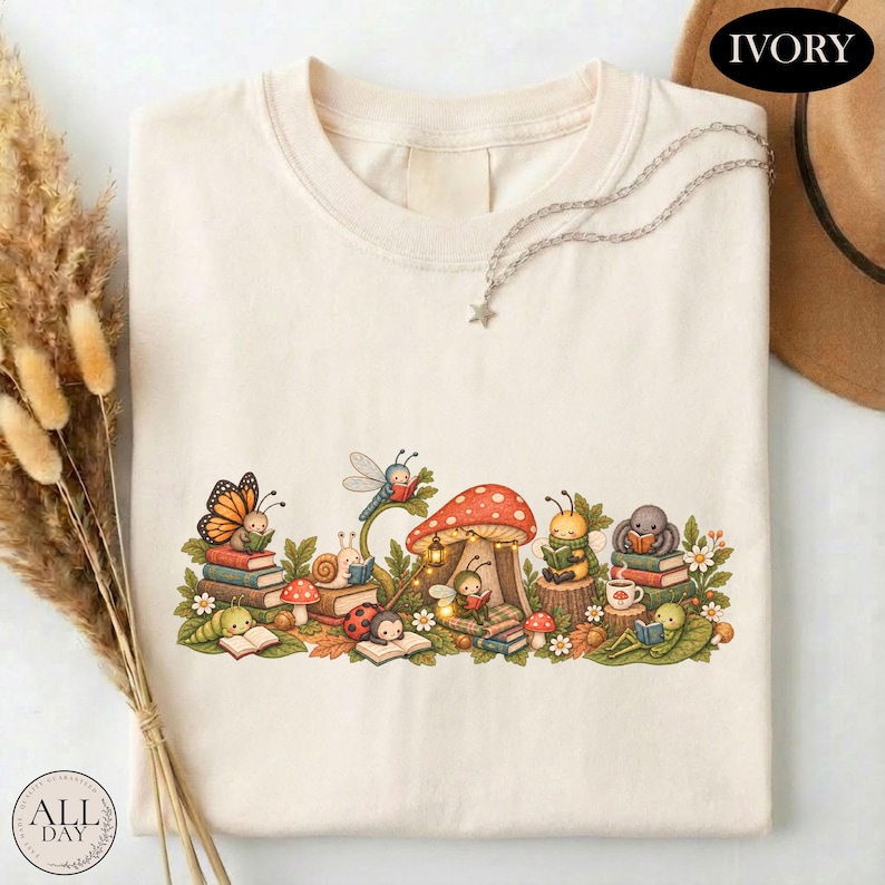 K&ouml;nnte beinhalten: Ein elfenbeinfarbenes T-Shirt mit einer grafischen Illustration verschiedener Insekten, darunter ein Schmetterling, eine Schnecke, ein Marienk&auml;fer und eine Biene, die um einen gro&szlig;en roten Pilz herum B&uuml;cher lesen. Das Design enth&auml;lt gr&uuml;ne Bl&auml;tter.