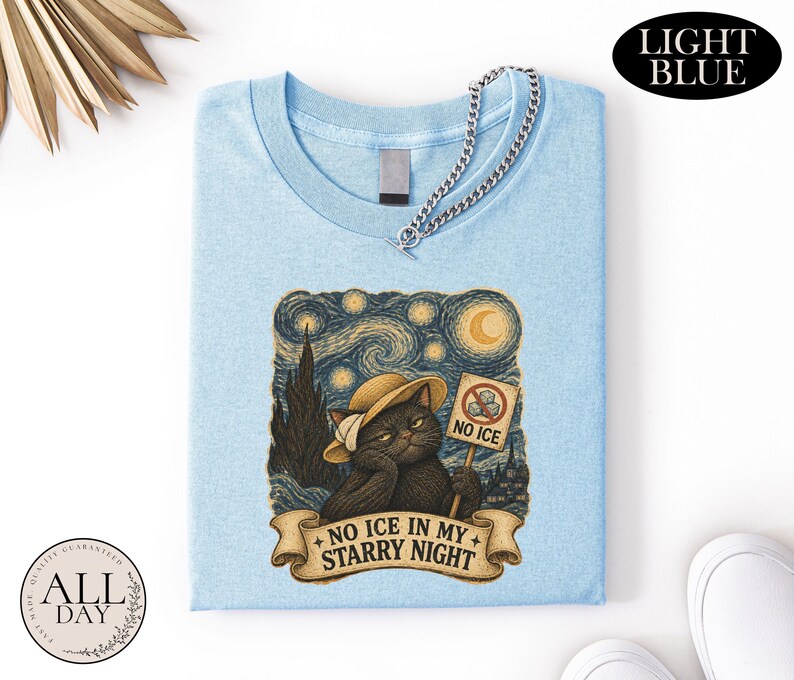 May include: A light blue t-shirt featuring a graphic of a black cat wearing a straw hat, holding a sign that says NO ICE with a crossed-out ice cube icon. Below the cat, a banner displays the text NO ICE IN MY STARRY NIGHT in dark lettering.