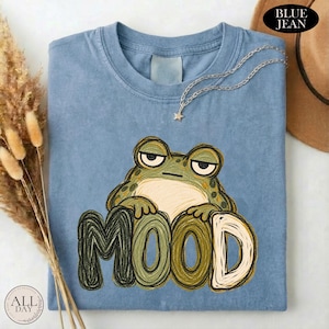 May include: A blue crew neck t-shirt featuring a graphic of a green frog with a bored expression resting its arms on the word MOOD written in large, textured, hand-drawn style letters. A delicate silver-tone chain necklace rests on the collar.