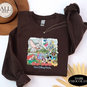 A brown t-shirt with a graphic of Disney characters from the Magic Kingdom, including Dumbo, Minnie Mouse, Mickey Mouse, Donald Duck, and other iconic figures, along with a castle and a tree. The t-shirt is displayed on a white background, and there is a brown hat and some yellow leaves in the foreground.