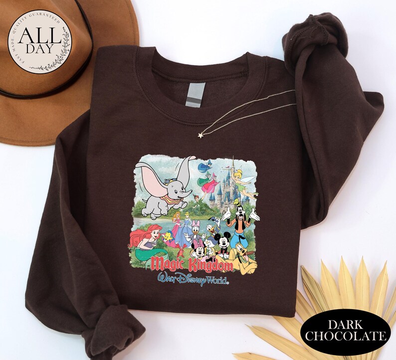 A brown t-shirt with a graphic of Disney characters from the Magic Kingdom, including Dumbo, Minnie Mouse, Mickey Mouse, Donald Duck, and other iconic figures, along with a castle and a tree. The t-shirt is displayed on a white background, and there is a brown hat and some yellow leaves in the foreground.