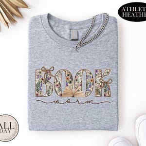 A gray t-shirt with the word "BOOK" printed on it, surrounded by floral designs. The t-shirt is folded and placed on a white surface, with a necklace and some decorative elements in the background.