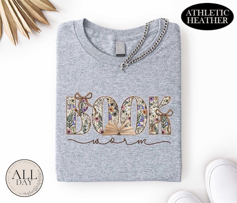 A gray t-shirt with the word "BOOK" printed on it, surrounded by floral designs. The t-shirt is folded and placed on a white surface, with a necklace and some decorative elements in the background.