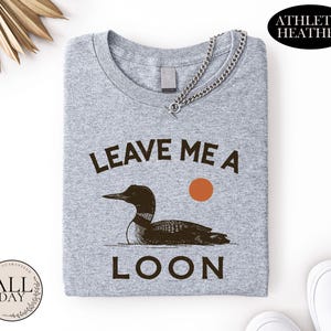 a gray t-shirt with a graphic design of a duck and the text "LEAVE ME A LON" printed on it.