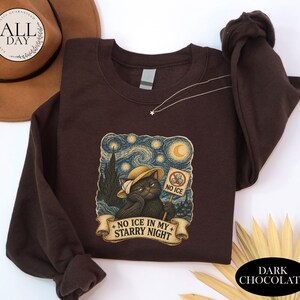 May include: A dark chocolate brown long-sleeve sweatshirt featuring a graphic print of a cat wearing a hat, holding a sign that says NO ICE, with the text NO ICE IN MY STARRY NIGHT on a banner below, set against a swirling starry night background.