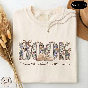 A white t-shirt with the word "BOOK" printed on it, surrounded by floral designs. The t-shirt is placed on a white surface, with some dried flowers and a hat visible in the background.