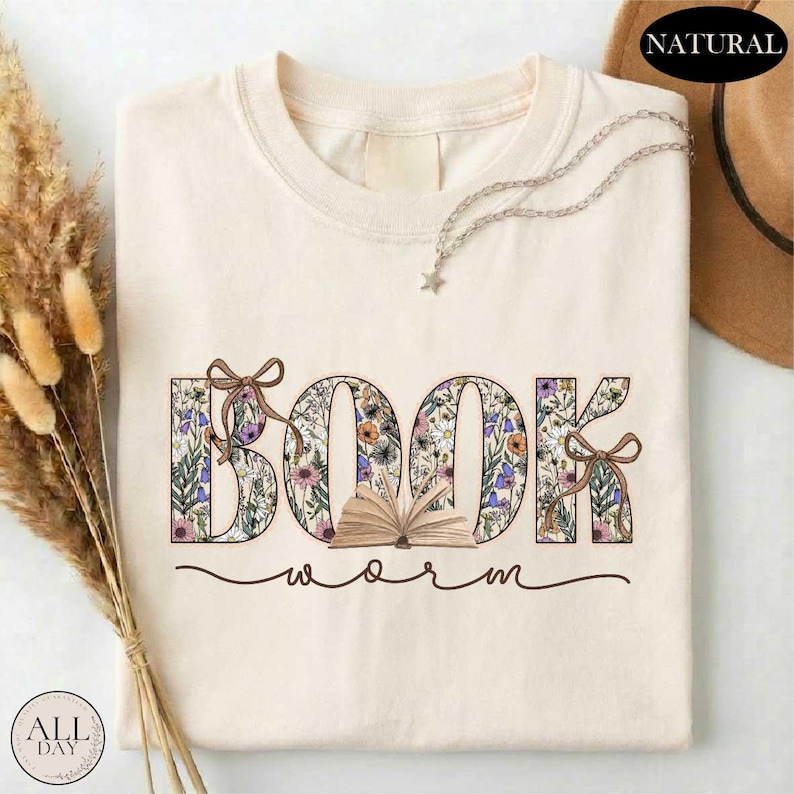 A white t-shirt with the word "BOOK" printed on it, surrounded by floral designs. The t-shirt is placed on a white surface, with some dried flowers and a hat visible in the background.