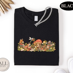 K&ouml;nnte beinhalten: Ein schwarzes T-Shirt mit einer grafischen Illustration verschiedener Insekten, darunter ein Schmetterling, eine Schnecke, ein Marienk&auml;fer und eine Biene, die um einen roten Pilz herum B&uuml;cher lesen. Eine silberfarbene Kette liegt am Halsausschnitt.
