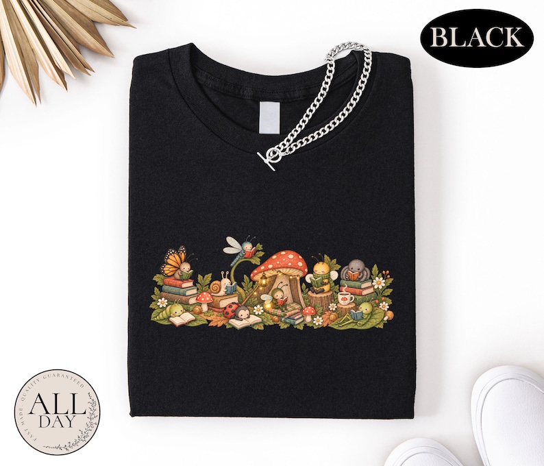 K&ouml;nnte beinhalten: Ein schwarzes T-Shirt mit einer grafischen Illustration verschiedener Insekten, darunter ein Schmetterling, eine Schnecke, ein Marienk&auml;fer und eine Biene, die um einen roten Pilz herum B&uuml;cher lesen. Eine silberfarbene Kette liegt am Halsausschnitt.