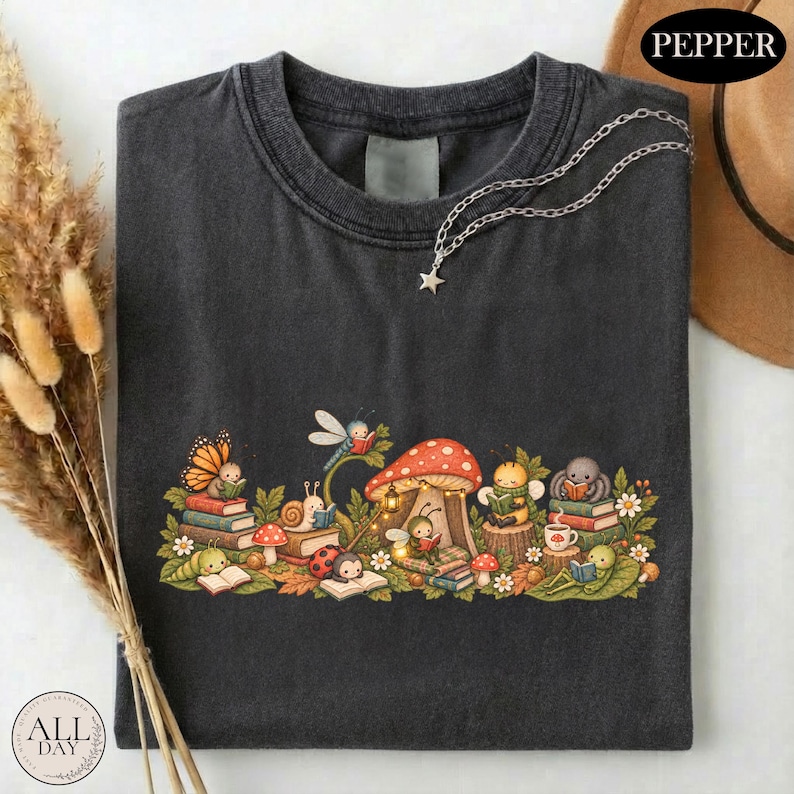 K&ouml;nnte beinhalten: Ein anthrazitfarbenes T-Shirt mit einer grafischen Illustration verschiedener Insekten, darunter ein Schmetterling, eine Schnecke, ein Marienk&auml;fer und eine Biene, die um einen gro&szlig;en roten Pilz herum B&uuml;cher lesen. Eine silberfarbene Kette mit Sternanh&auml;nger liegt am Kragen.