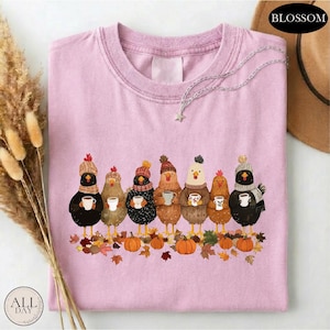 May include: A light pink crew neck t-shirt featuring a graphic of seven chickens wearing knit hats and scarves, each holding a white mug. The chickens stand above a pile of autumn leaves and orange pumpkins. A silver chain necklace rests on the collar.