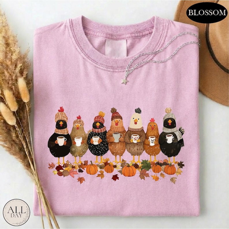 May include: A light pink crew neck t-shirt featuring a graphic of seven chickens wearing knit hats and scarves, each holding a white mug. The chickens stand above a pile of autumn leaves and orange pumpkins. A silver chain necklace rests on the collar.