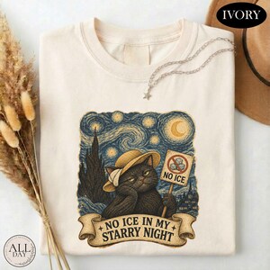 May include: An ivory t-shirt featuring a graphic of a black cat wearing a straw hat against a swirling blue starry night background. The cat holds a sign with a crossed-out ice cube and the text NO ICE. Below, a banner reads NO ICE IN MY STARRY NIGHT.