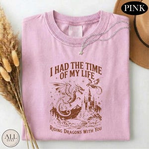 A pink t-shirt with a graphic design of a dragon and castle, along with the text "I HAD THE TIME OF MY LIFE RIDING DRagons WITH YOU".
