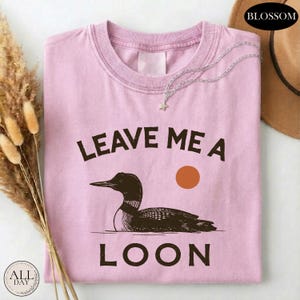 A pink t-shirt with a graphic design of a duck and the text "LEAVE ME A LON" printed on it, along with a small star and a sun graphic.