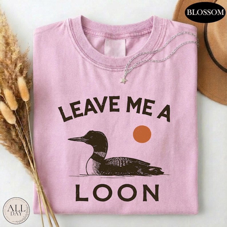A pink t-shirt with a graphic design of a duck and the text "LEAVE ME A LON" printed on it, along with a small star and a sun graphic.
