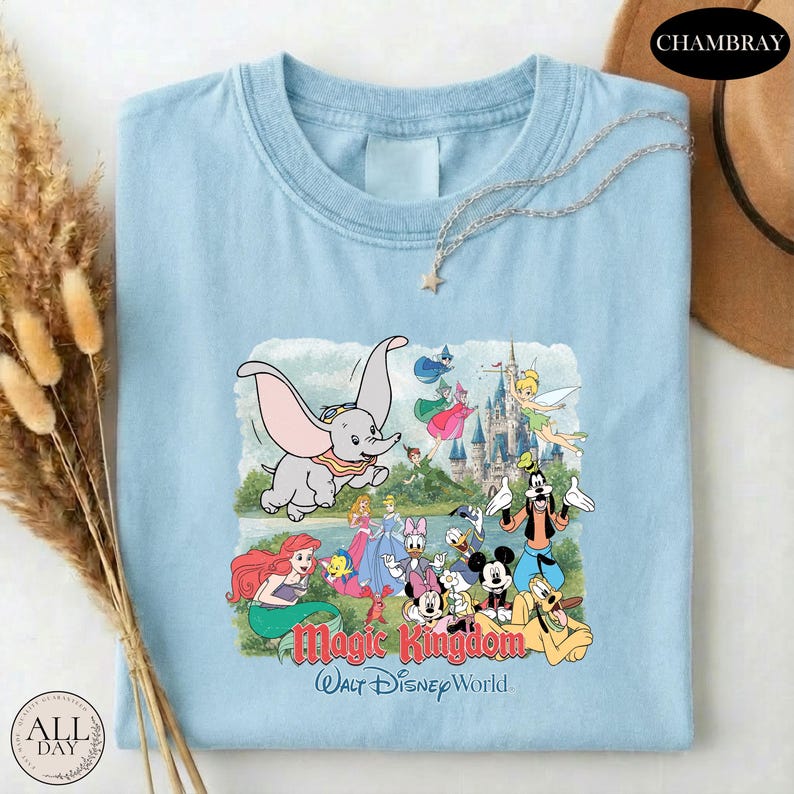 A light blue t-shirt with a Disney-themed graphic of characters from the Magic Kingdom, including Dumbo, Minnie Mouse, Mickey Mouse, Donald Duck, and other iconic Disney characters, along with a castle and a star.