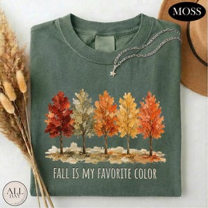May include: A moss green crew neck t-shirt featuring a graphic of five trees in autumn colors including red, orange, and yellow. Below the trees, the text reads "FALL IS MY FAVORITE COLOR" in white capital letters. A silver star charm necklace is included.