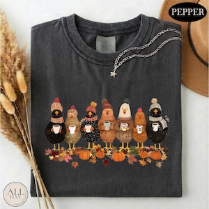 May include: A charcoal gray t-shirt featuring a graphic of seven chickens wearing knit hats and scarves, each holding a white mug. The chickens stand on a pile of autumn leaves and pumpkins. A silver star-shaped pendant hangs from the neckline.