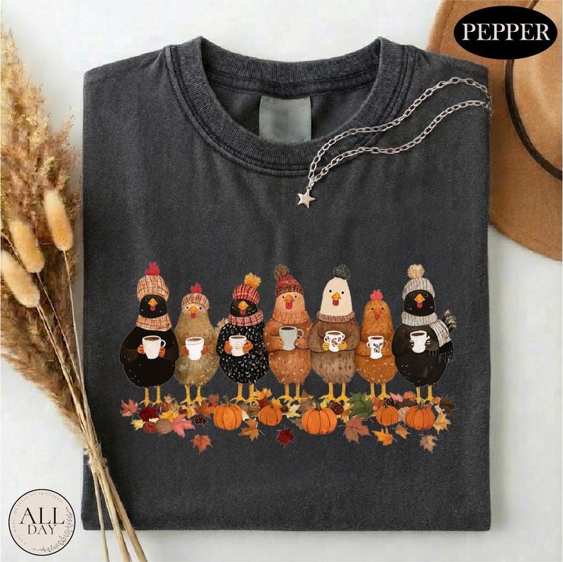 May include: A charcoal gray t-shirt featuring a graphic of seven chickens wearing knit hats and scarves, each holding a white mug. The chickens stand on a pile of autumn leaves and pumpkins. A silver star-shaped pendant hangs from the neckline.
