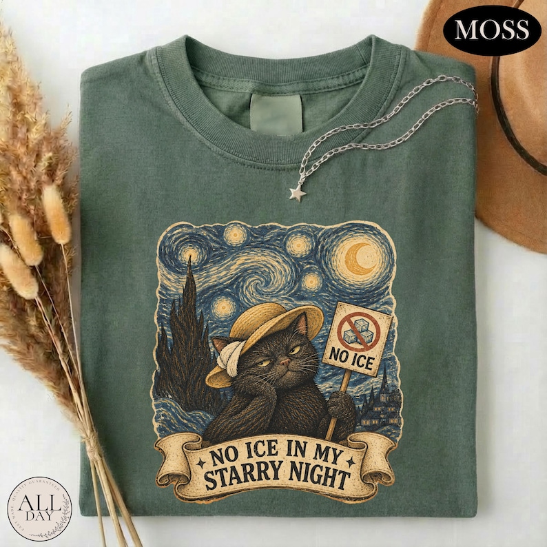 May include: A sage green t-shirt featuring a graphic of a black cat wearing a straw hat, holding a sign that says NO ICE with a crossed-out ice cube icon. Below the cat, a banner reads NO ICE IN MY STARRY NIGHT. A silver star necklace rests on the collar.