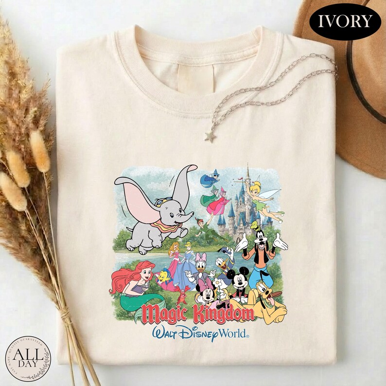 A white t-shirt with a colorful graphic of Disney characters from the Magic Kingdom, including elephants, fairies, and other iconic figures, along with the text "Magic Kingdom" and "Walt Disney World."