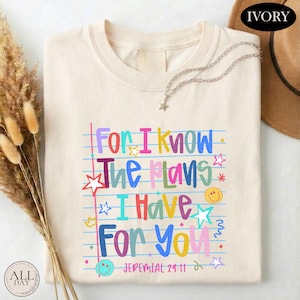 May include: An ivory t-shirt featuring a graphic print of blue horizontal lines resembling notebook paper. The text reads "For I know The plans I Have For You" in multi-colored letters, accompanied by small star and smiley face icons.