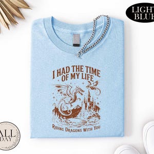 a light blue t-shirt with a graphic design depicting a dragon and castle, along with the text "I HAD THE TIME OF MY LIFE RIDING DRagons WITH YOU". The t-shirt is folded and displayed on a white background, with a small circular logo in the bottom left corner that reads "ALL DAY".