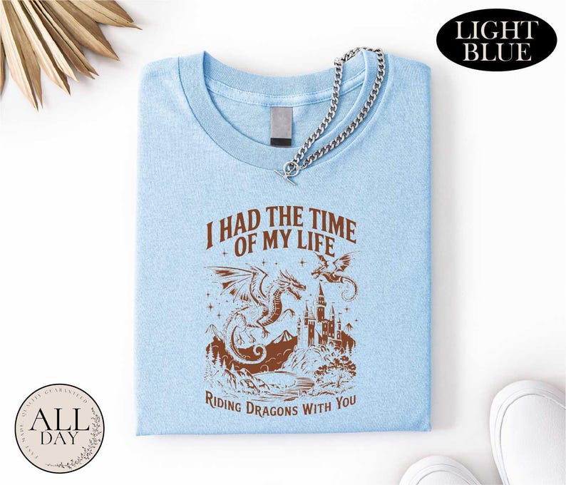 a light blue t-shirt with a graphic design depicting a dragon and castle, along with the text "I HAD THE TIME OF MY LIFE RIDING DRagons WITH YOU". The t-shirt is folded and displayed on a white background, with a small circular logo in the bottom left corner that reads "ALL DAY".