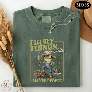 May include: A moss green t-shirt featuring a graphic of a frog wearing a straw hat and blue overalls, holding a shovel. The text reads I BURY THINGS... may be plants... MAYBE PEOPLE. A silver star-shaped necklace rests on the collar of the shirt.