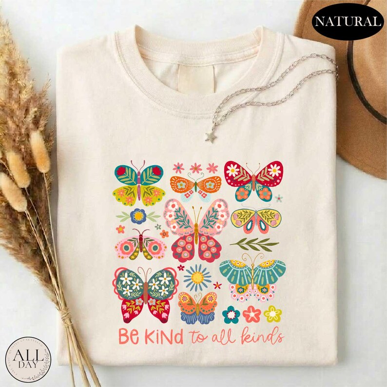 A white t-shirt with a colorful graphic design of butterflies and flowers, along with the text "Be Kind to all kinds" printed on it.