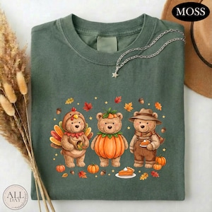 May include: A dark olive green t-shirt featuring a graphic of three bears dressed for autumn. One bear wears a turkey costume, one is inside a pumpkin, and one wears overalls and a hat. The design includes small pumpkins, acorns, and leaves.