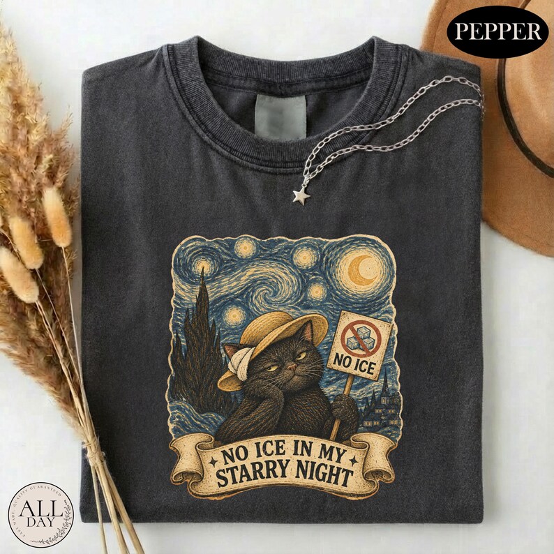 May include: A charcoal gray t-shirt featuring a graphic of a black cat wearing a straw hat, set against a swirling starry night background. The cat holds a sign that reads NO ICE, and a banner below displays the text NO ICE IN MY STARRY NIGHT.
