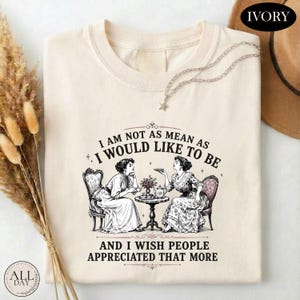 May include: An ivory t-shirt featuring a graphic of two women sitting at a small table with tea, accompanied by the text: I AM NOT AS MEAN AS I WOULD LIKE TO BE AND I WISH PEOPLE APPRECIATED THAT MORE. The design is rendered in a dark brown ink.