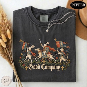 May include: A charcoal gray t-shirt featuring a graphic of two rabbits and a cat dressed in medieval attire with capes and shields. One rabbit plays a trumpet, another rides a dog, and the cat carries a flag. The text Good Company is printed below.