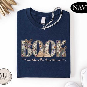 A navy blue t-shirt with the word "BOOK" embroidered on it, along with the word "Warm" written below it. The t-shirt is folded and displayed on a white background, with a necklace and some decorative elements visible in the top corners.