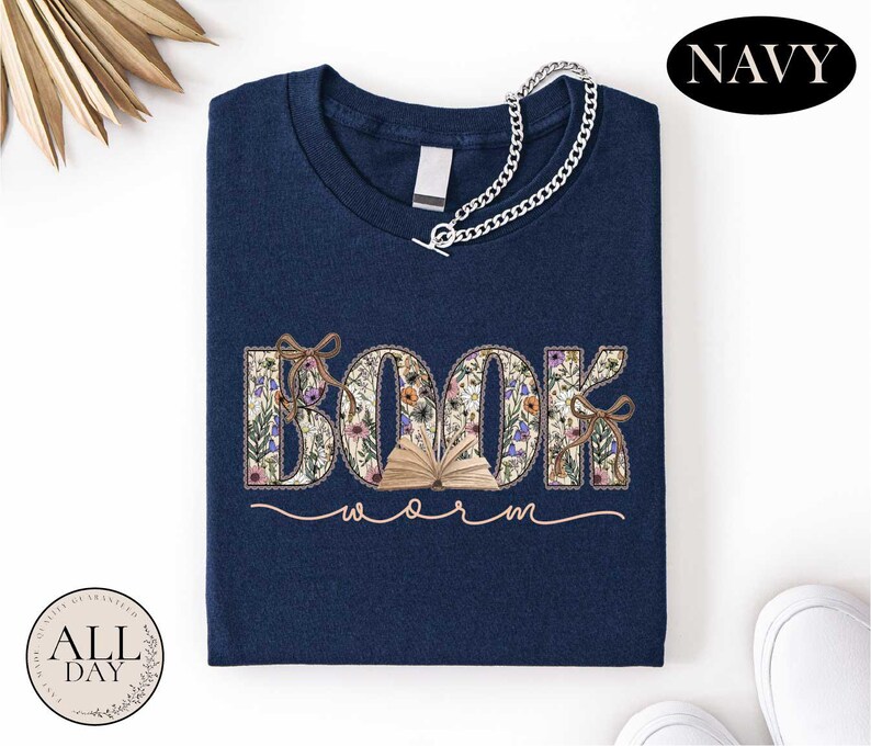 A navy blue t-shirt with the word "BOOK" embroidered on it, along with the word "Warm" written below it. The t-shirt is folded and displayed on a white background, with a necklace and some decorative elements visible in the top corners.