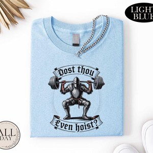 a light blue t-shirt with a graphic design depicting a knight lifting a barbell, accompanied by the text "Post thou Even hoist?"