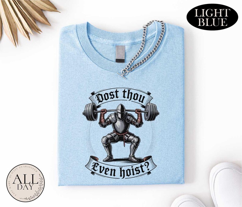 a light blue t-shirt with a graphic design depicting a knight lifting a barbell, accompanied by the text "Post thou Even hoist?"