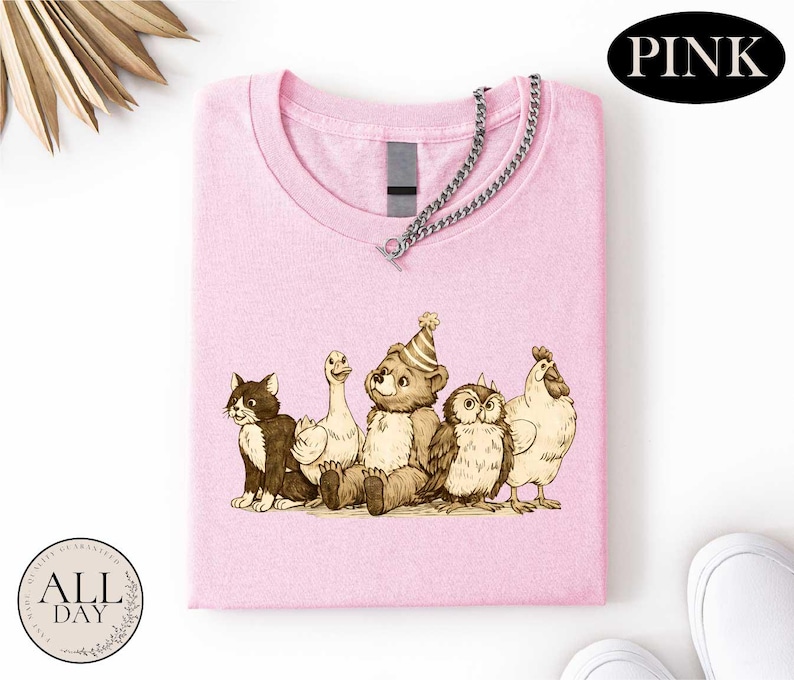 A pink t-shirt with a cartoon illustration of a bear, cat, duck, chicken, and owl wearing party hats.