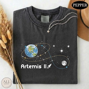 May include: Dark gray t-shirt featuring a graphic of Earth, a rocket, and the moon, with a star-shaped necklace. The text "Artemis II" is printed in white. The shirt has a crew neck and is folded.