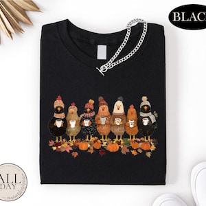 May include: A black t-shirt featuring a graphic of seven chickens wearing colorful knit hats and scarves, each holding a white mug. The chickens stand on a pile of autumn leaves and orange pumpkins. A silver chain is attached to the neckline.