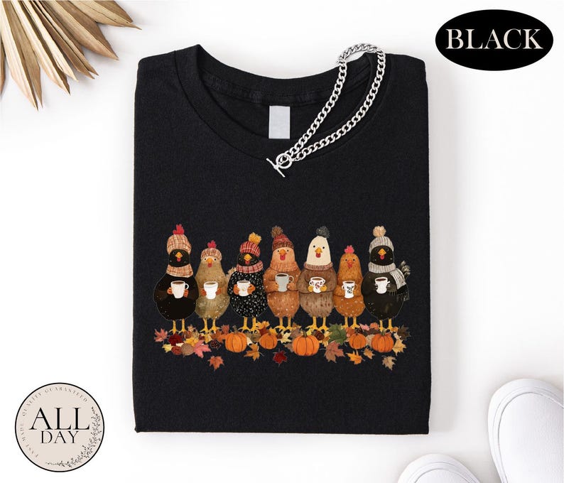 May include: A black t-shirt featuring a graphic of seven chickens wearing colorful knit hats and scarves, each holding a white mug. The chickens stand on a pile of autumn leaves and orange pumpkins. A silver chain is attached to the neckline.