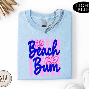 a light blue t-shirt with the text "Beach Bum" printed on it, along with pink and purple floral designs. The t-shirt is folded and placed on a white surface, with a chain visible around the neck. There are also some other items in the image, such as a pair of white shoes and a small circular object.