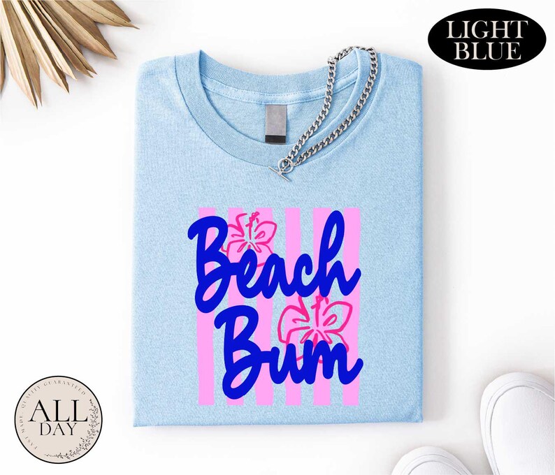 a light blue t-shirt with the text "Beach Bum" printed on it, along with pink and purple floral designs. The t-shirt is folded and placed on a white surface, with a chain visible around the neck. There are also some other items in the image, such as a pair of white shoes and a small circular object.