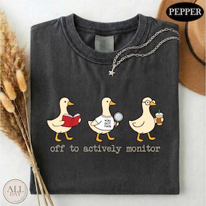 May include: Dark gray t-shirt featuring three cartoon ducks. One duck reads a book, another holds a magnifying glass, and the third has glasses and a coffee cup. The text "off to actively monitor" is printed below the ducks.